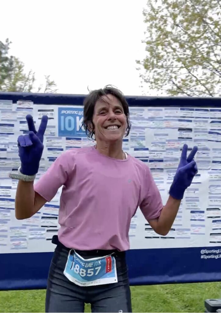 Smiling runner in pink shirt making peace signs after a 10K race.